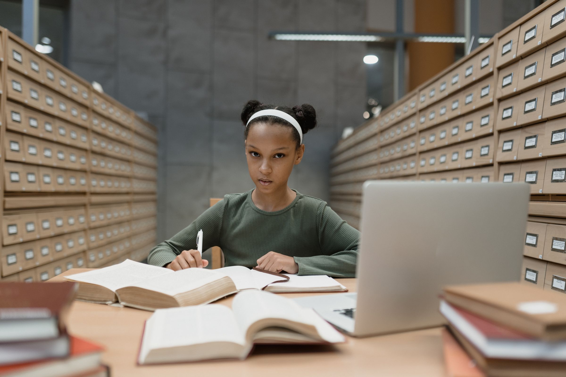 A student writing notes at a desk with open books and a laptop, between rows of library card catalogues